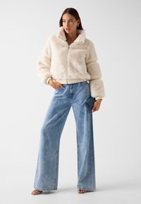 Kremowa kurtka z imitacji futra z wysokim kołnierzem, zamkiem błyskawicznym i prążkowanym dołem. Połączona z jasnoniebieskimi, szerokimi spodniami i małą, ciemną kopertówką.