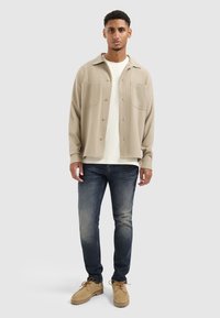 Beige knoop-overhemd met twee voortassen, gedragen over een wit T-shirt, gecombineerd met donkere jeans en tan schoenen. Neutraal kleurenpalet.