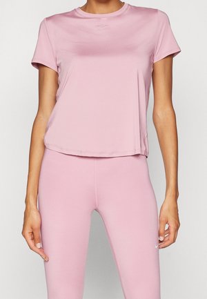 Vrouw die een aansluitend lichtroze sportshirt met korte mouwen en bijpassende legging draagt, staand tegen een effen achtergrond.