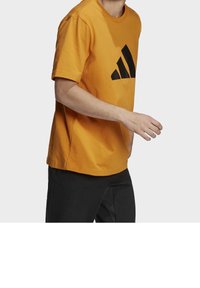 Orange bomullströja med en stor svart Adidas-logotyp på framsidan. Tröjan har korta ärmar och rund halsringning, i kombination med svarta byxor.