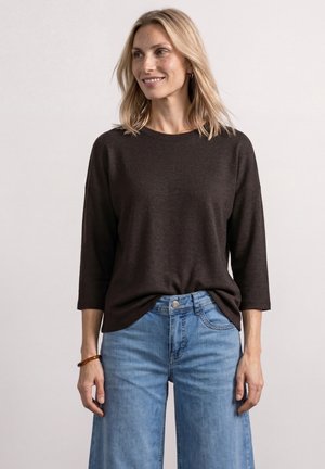 Femme blonde portant un haut marron foncé à manches trois-quarts et un jean taille haute bleu clair, debout devant un fond uni.