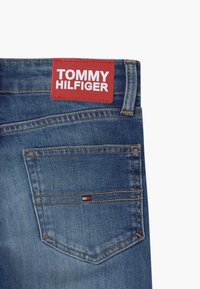 Modré džíny z denimu s červenou koženou visačkou s embosovaným nápisem "TOMMY HILFIGER" v bílé barvě. Na zadní kapse se nachází malá vícebarevná nášivka.