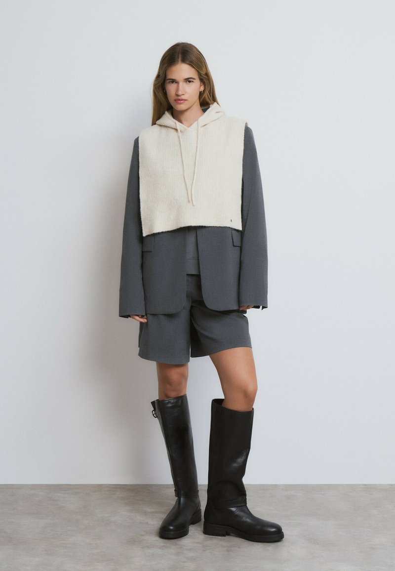 Grau blazer über passenden Shorts, einen cremefarbenen Cropped-Hoodie und schwarze kniehohe Lederstiefel mit glatter Oberfläche und minimalen Details.