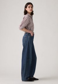 Chemise à col rayée rouge et blanche, jean large taille haute bleu, et chaussures plates noires. Le denim présente des coutures visibles et des poches.