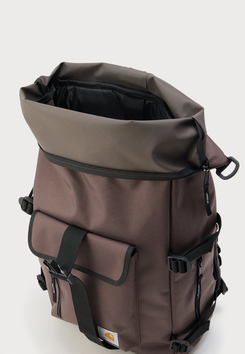 Sac à dos marron à rabat avec poche avant à boucle, sangles latérales et compartiment principal partiellement ouvert montrant la doublure intérieure noire.