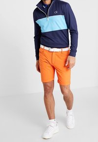 Námořnická a světle modrá mikina na zip s vysokým límcem, doplněná oranžovými šortkami. Bílý opasek a tenisky dotvářejí outfit. Hladké textury.