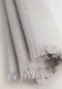 Tissu tissé gris clair avec bords effrangés et une ligne de petits sequins transparents le long du pli, suspendu en plis doux.