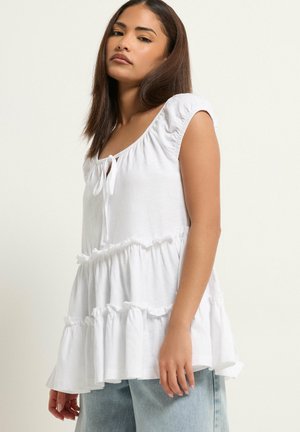 Junge Frau mit glattem braunem Haar trägt eine weiße, ärmellose Bluse mit Rüschen und hellblaue Jeans, sie steht vor einem schlichten Hintergrund.