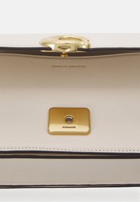 Borsa a tracolla in pelle beige con chiusura oro, rifiniture nere e decorazione con marchio in metallo dorato. Interno con texture morbida e stampa del logo.