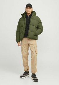 Puffjakke i mørkegrøn, lynlås med hætte. Sort fleece underneath, beige cargo-bukser og sorte sneakers med grå accenter.