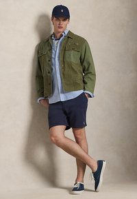 Mladý muž nosiaci námornícku čiapku, olivovo zelenú bundu, svetlomodrú košeľu, námornícke šortky a námornícke tenisky, stojaci pred jednoduchou béžovou stenou.