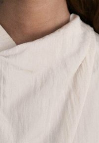 Tessuto bianco, texturizzato, con una morbida caduta e un colletto arricciato, che mette in mostra un aspetto naturale, non sbiancato, e dettagli di design minimal.
