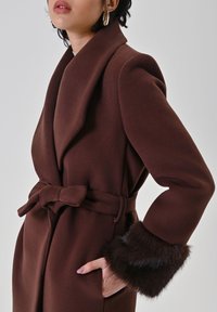 Cappotto marrone con cintura e ampi revers, caratterizzato da polsini bordati di pelliccia. Realizzato in tessuto morbido con una texture liscia, presenta una silhouette sartoriale.