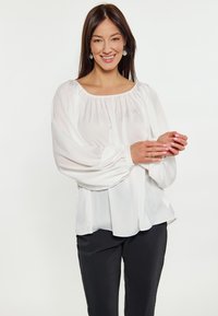 Weiße, leichte Bluse mit elastischem Halsausschnitt und weit ausgestellten Ärmeln, mit einer weichen, glatten Textur. Kombiniert mit schwarzen Hosen.