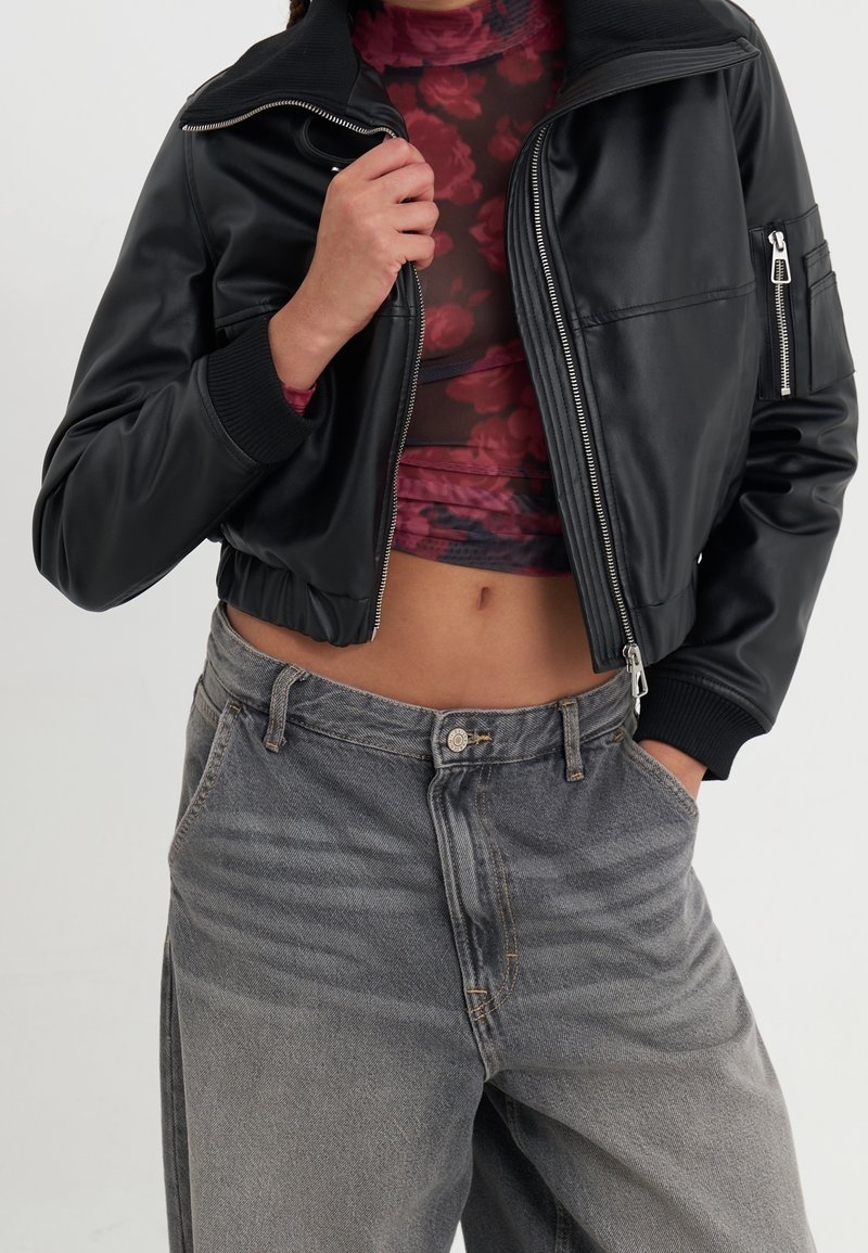 Chaqueta bomber de cuero sintético negro con puños acanalados y bolsillos con cremallera, combinada con jeans de tiro alto de color gris ligeramente desgastados.