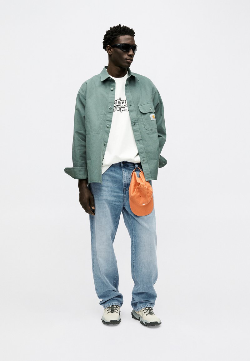 Man wearing black sunglasses, green overshirt, white graphic t-shirt, blue jeans, and holding orange cap clipped to belt loop.