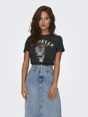 Jonge vrouw met krullend haar, gekleed in een zwart t-shirt met een grafische print van luipaardmotief en een spijkerrok met hoge taille, staand tegen een effen achtergrond.