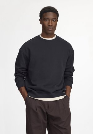 Giovane uomo che indossa una felpa blu scuro sopra una camicia chiara, con le mani nelle tasche, in piedi davanti a uno sfondo bianco semplice.
