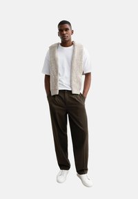 Un gilet en tricot beige clair par-dessus un t-shirt blanc, associé à un pantalon marron foncé et des baskets blanches, créant une tenue en couches.