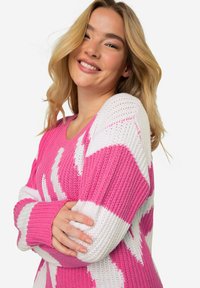 Rosa och vit stickad tröja med ett strukturerat mönster, som har långa ärmar och en vid halsringning. Designen inkluderar djärva zigzag-ränder.