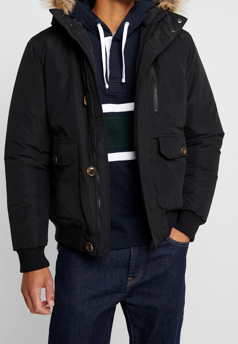 Sort dunjakke med pelsforet hætte, lynlås og knaplukning, forlommer, ribstrikkede ærmeafslutninger, båret over en marineblå hættetrøje med striber.