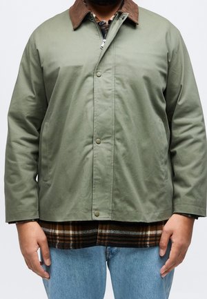 Homme portant une veste vert clair avec un col marron, une chemise à carreaux en dessous, et un jean bleu clair, debout avec les mains détendues le long du corps.