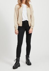 Frau trägt eine beigefarbene Bomberjacke, ein weißes T-Shirt, schwarze Skinny Jeans und schwarze Schnürstiefel mit Plateausohle, steht vor einem weißen Hintergrund.