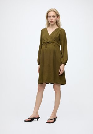 Femme portant une robe de maternité vert olive à manches longues et des sandales noires à talon bas, debout devant un fond blanc uni.