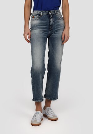 Jean en denim bleu délavé avec une coupe raccourcie, jambe droite et ourlet effiloché. Portés avec des baskets gris clair dotées de lacets blancs.
