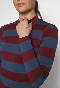 Langermet topp med vekselvis maroon og blå horisontale striper. Stoffet er glatt, med en tettsittende design og høy krage.