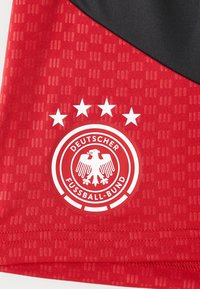 Κόκκινη αθλητική φανέλα με υφή. Εμφανίζει το λογότυπο �της Deutscher Fußball-Bund με έναν αετό και τέσσερα αστέρια από πάνω.