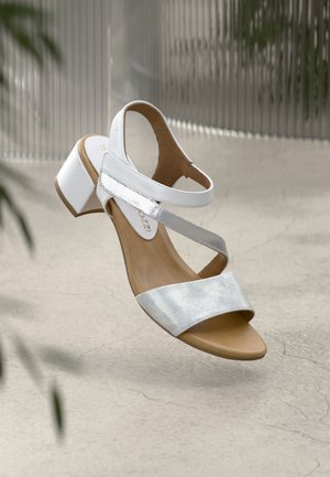 Άσπρο σανδάλι με ασημένιο μεταλλικό λουρί, ανοιχτό δάχτυλο και μέτριο τακούνι. Λείο υλικό με εσωτερική σόλα σε ανοικτό καφέ χρώμα. Ρυθμιζόμενο λουρί στον αστράγαλο.