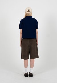 Personne portant une casquette beige, une chemise à manches courtes bleu marine, un short marron jusqu'aux genoux, des chaussettes blanches montantes et des chaussures foncées, debout de dos.