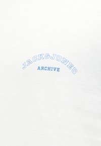 Λευκό ύφασμα με ανοιχτό μπλε κείμενο "JACK & JONES" σε καμάρα πάνω από τη μικρότερη λέξη "ARCHIVE" στο κέντρο της επιφάνειας.