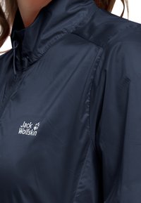 Mörkblå lättviktsjacka med hög krage och dragkedja, med slät textur och en vit logotyp på bröstet.