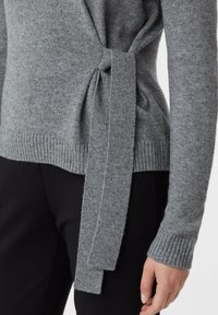 Nahaufnahme einer Person, die einen grauen Strickpullover mit seitlicher Bindung und schwarze Hose trägt, wobei Arm und Hand zu sehen sind.