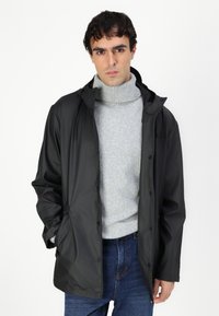 Veste noire imperméable avec capuche, fermeture à boutons et deux poches latérales, portée sur un pull gris clair et un jean bleu.