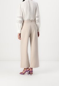 Vaaleanbeige leveät housut yhdistettynä valkoiseen paitaan, jossa on pitsiyksityiskohtia hihoissa ja selässä. Mallilla on päällään violetit nahanauha-korkokengät.