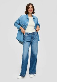Helles, blauschattiertes Denim-Jacket über einem weißen Oberteil, kombiniert mit hellblauen weiten Jeans. Weiße Sneakers und ein neutraler Hintergrund runden den Look ab.