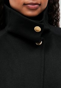 Person iført en sort frakke med høj krave, der er fastgjort med to guldknapper, der viser den nederste del af deres ansigt og krøllet hår.