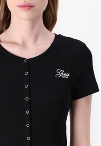 Musta ribbattu t-paita pyöreällä kaula-aukolla, jossa on hopeaiset napit ja valkoinen brodeerattu "Guess Jeans" -logo rinnassa.