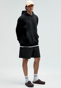 Zwarte capuchontrui met voorzak, gecombineerd met zwarte shorts. Licht perzikkleurige pet, witte sokken met grijze strepen en bruine klompen.