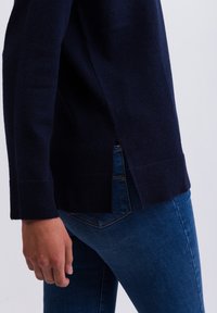 Pull en tricot bleu marine avec des poignets côtelés, des fentes latérales et une coupe décontractée, associé à un jean en denim ajusté. La texture douce est visible.