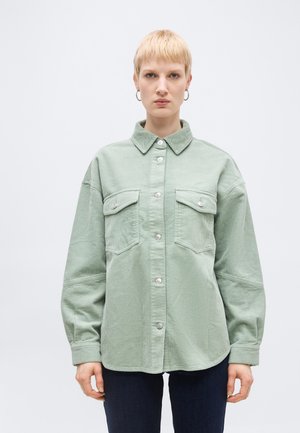 Person with short blonde hair and hoop earrings wearing a light green corduroy button-up shirt with chest pockets, standing against a plain background.