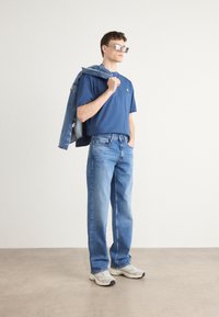 Blå t-shirt med et lille logo, lyseblå denimjeans og hvide sneakers. En denimjakke er draperet over den ene skulder. Firkantede solbriller bæres.