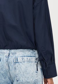 Navy blue long-sleeve shirt with a gathered hem, paired with light blue distressed denim jeans. Shirt has adjustable cuffs and smooth texture.