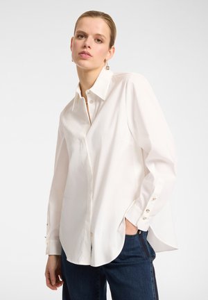 Chemise blanche à manches longues avec un col boutonné, coupe ample et ourlet arrondi. Boutons dorés aux poignets. Portée avec un denim foncé.