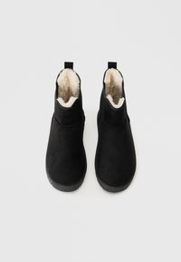 Juodi kulkšniniai batai su smėlio spalvos pūkų pamušalu, suapvalintais pirštais ir kilpomis, rodomi šalia vienas kito baltame fone.