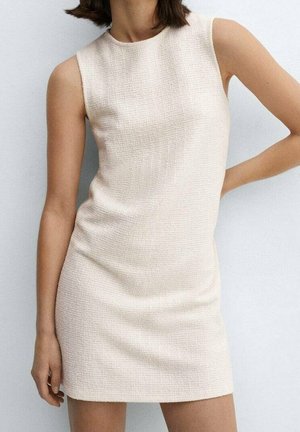 Ärmelloses, strukturiertes, beiges Minikleid, getragen von einer Frau, die eine Hand in die Hüfte gestützt hat und die andere herabhängt.