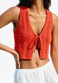 Rood gebreid cropped topje met een V-hals, voorzien van een getextureerd kabelpatroon en een striksluiting aan de voorkant, gecombineerd met witte broek met elastische tailleband.